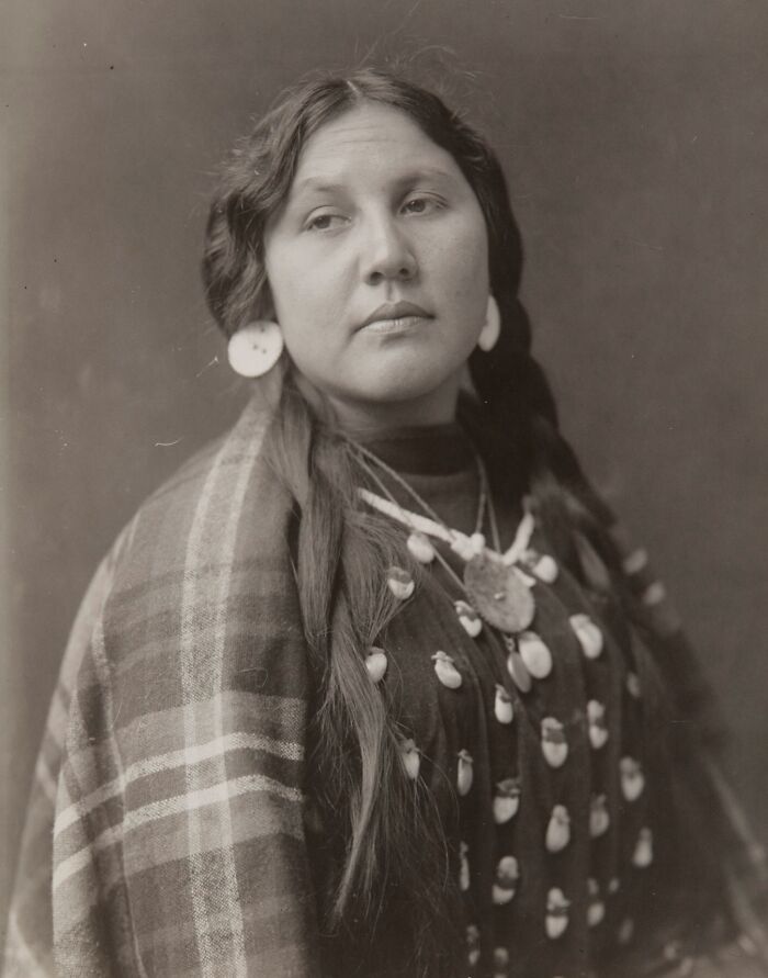 Native American woman from a century ago wearing traditional jewelry and a plaid shawl in a haunting black and white portrait.