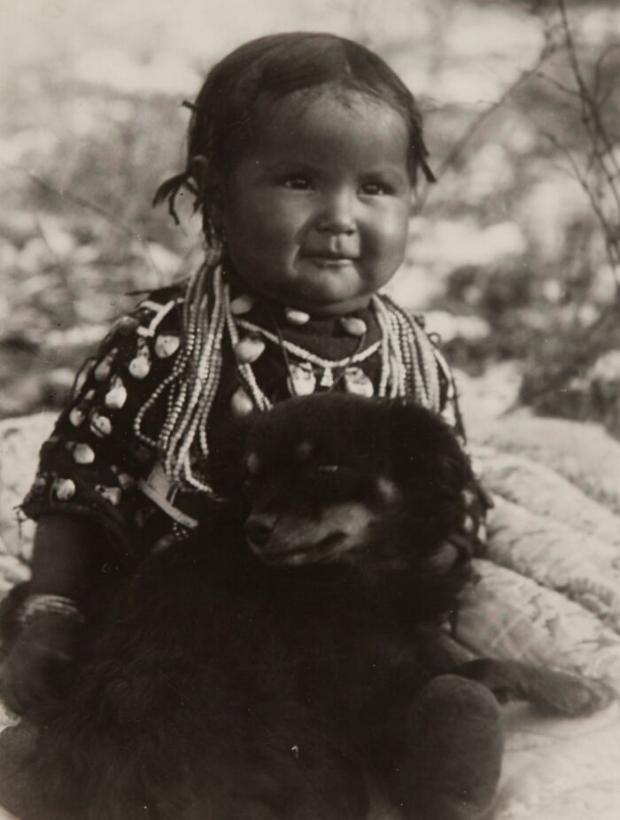 Native American child dressed in traditional beadwork, sitting with a dark dog in a vintage black and white photo.