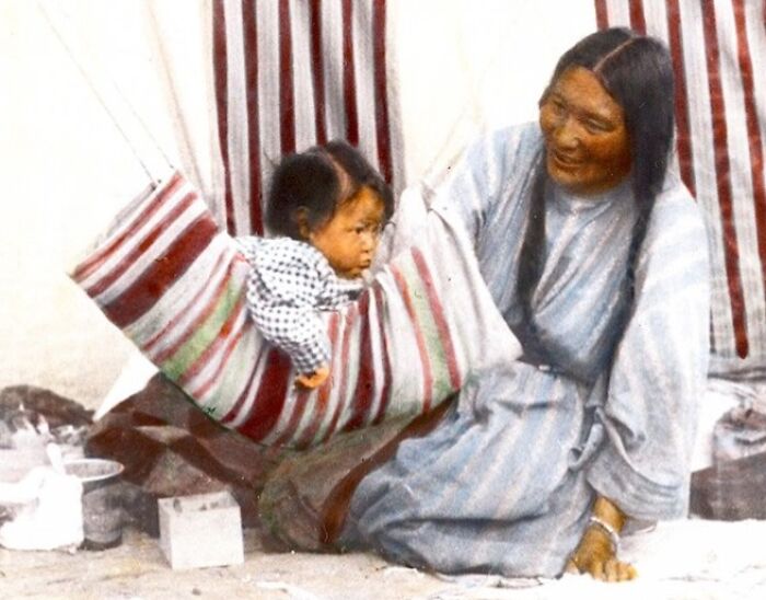 Native American woman smiling while holding a child in a traditional cradleboard wrapped with striped fabric.