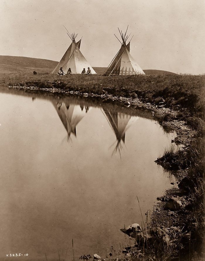 Two Native American teepees and people sitting by a riverbank, reflecting real lives from a century ago.