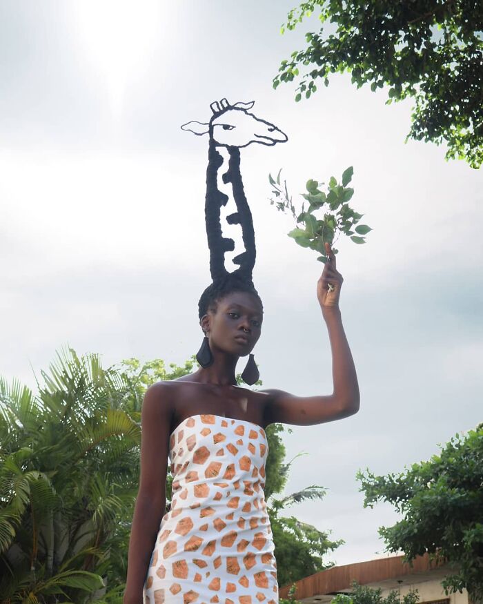 Woman posing with hair styled into a giraffe sculpture, holding leaves under a partly cloudy sky.