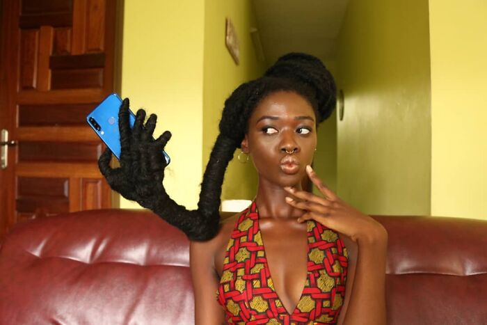 Artist with hair sculpture styled as a hand holding a phone, sitting on a red couch, wearing a patterned top.