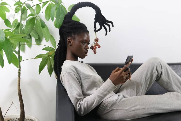 Woman with hair sculpture resembling grapes, relaxing on couch, holding a phone, next to a green plant.