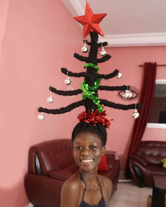 Woman with hair styled into a Christmas tree sculpture, featuring ornaments and a star, in a living room setting.