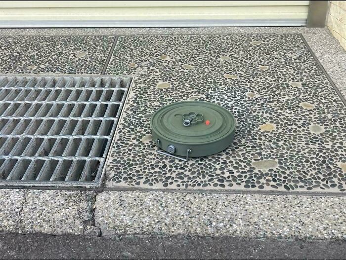 An unremarkable photo of a metal grate and a green round object on a speckled concrete surface outdoors.