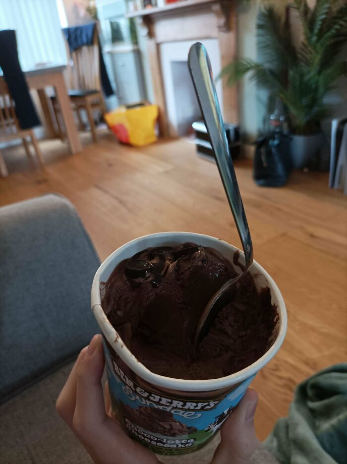Hand holding a spoon in a tub of chocolate ice cream in a living room, an unremarkable photo but entertaining.