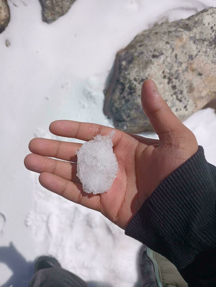 Hand holding a small chunk of snow outdoors with rocks and snow background in an unremarkable photo.