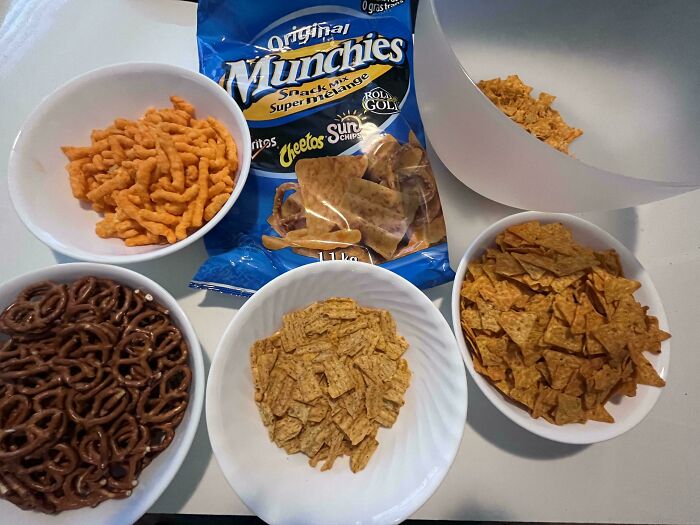 Various salty snacks including pretzels, cheesy puffs, and chips in bowls next to an original Munchies snack mix bag.