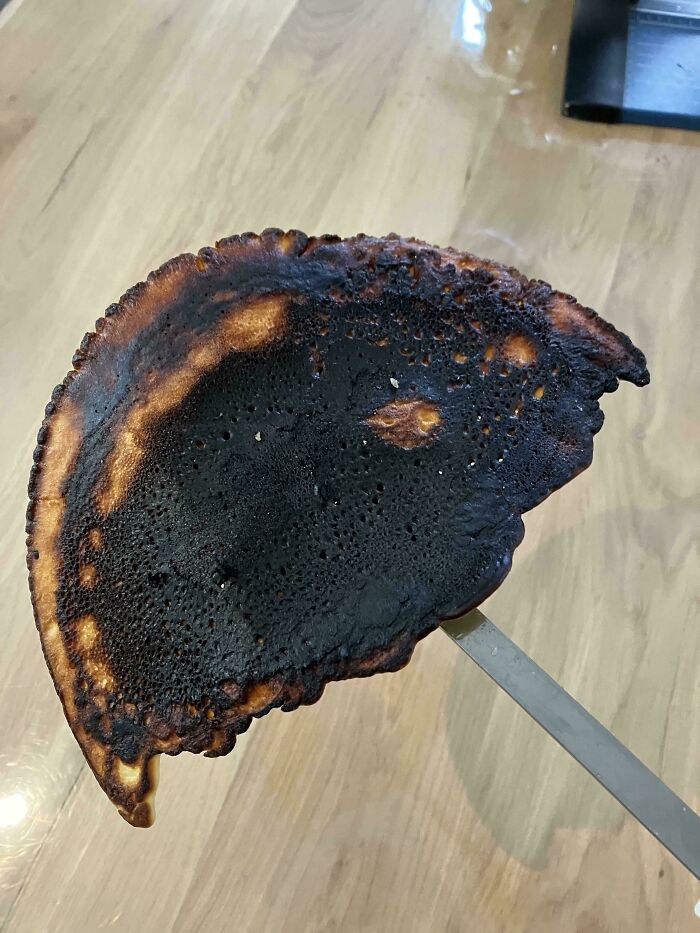 Burnt pancake held on a spatula showing a blackened surface in a kitchen, an unremarkable photo entertaining viewers.
