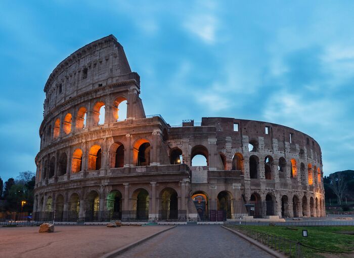 Ancient man-made structure of the Colosseum in Rome illuminated at dusk with a cloudy blue sky background.