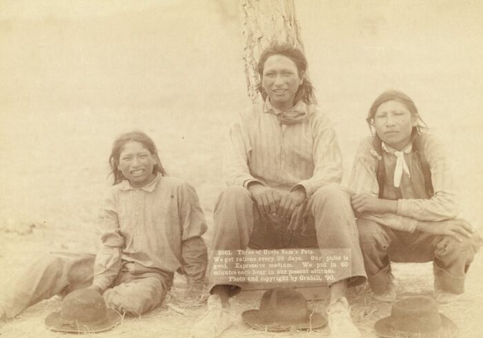 Three Native American men sitting outdoors in the 1800s, wearing period clothing and hats nearby, authentic photo.