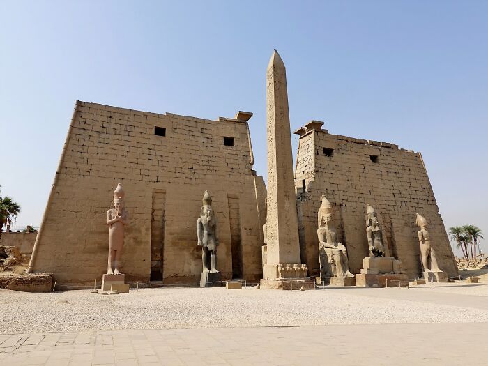 Ancient man-made structures including statues and an obelisk at an Egyptian temple complex under a clear sky.