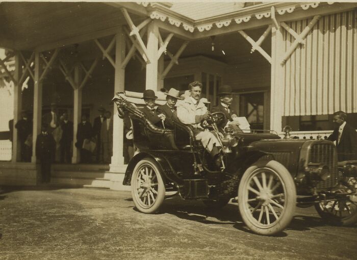 Early 1900s photo showing life in America with vintage car and people dressed in period clothing near a building.