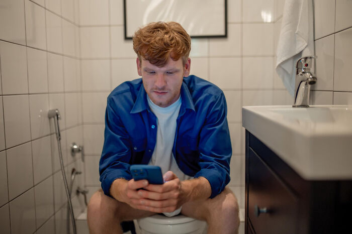 Man in blue shirt sitting on toilet using smartphone, capturing dating dynamics humor.