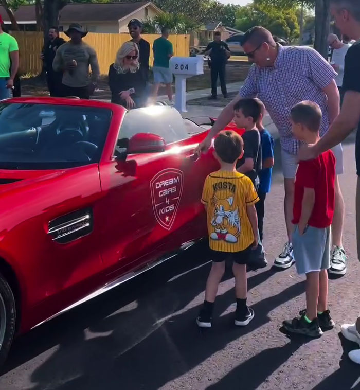 Children gathered around a red sports car with "Dream Cars 4 Kids" logo during a birthday surprise. Children gathered around a red sports car with "Dream Cars 4 Kids" logo during a birthday surprise.