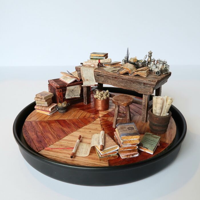 Miniature scene by Tamar Cohen depicting a detailed tiny desk with books, papers, and a stool, showcasing intricate craftsmanship.