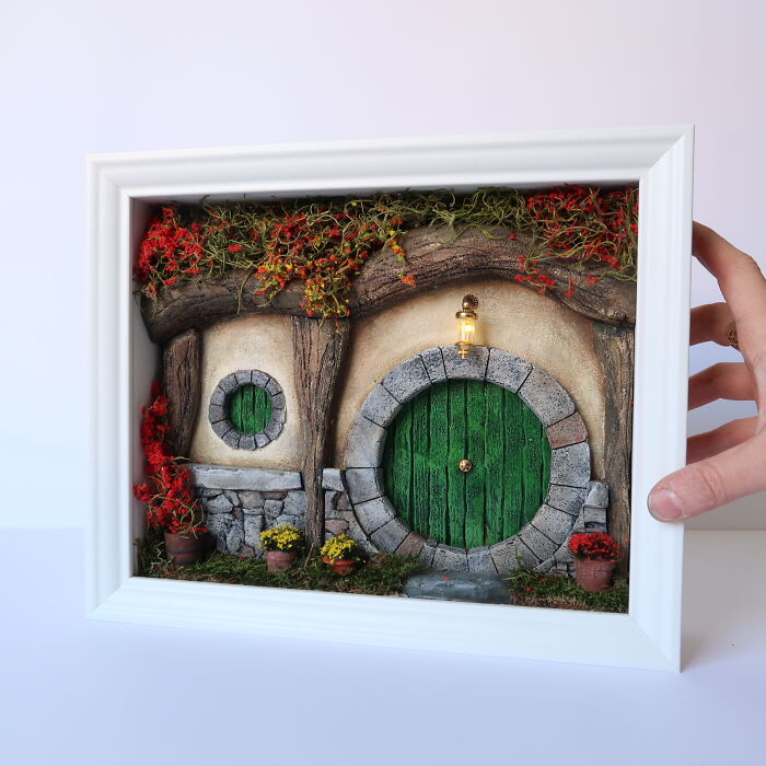 Miniature hobbit home by Tamar Cohen, featuring a green door and vibrant plants, held in a white frame.