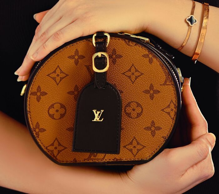 Designer bag held by hands, featuring gold accents and monogram pattern, symbolizing luxury pretensions.