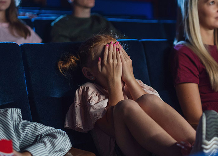 Child in theater covering face, reacting to event manager being humiliated on stage, seated in dimly lit audience area. Child in theater covering face, reacting to event manager being humiliated on stage, seated in dimly lit audience area.