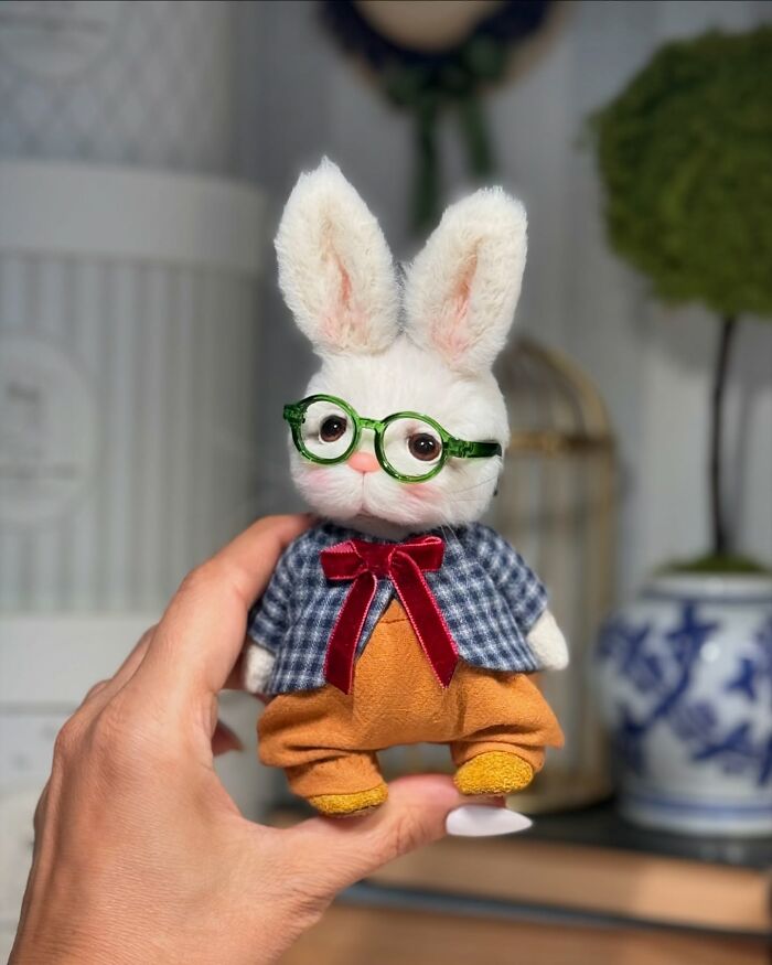 Handmade plush toy bunny with glasses, dressed in a blue checkered jacket and red bow, held in a hand.