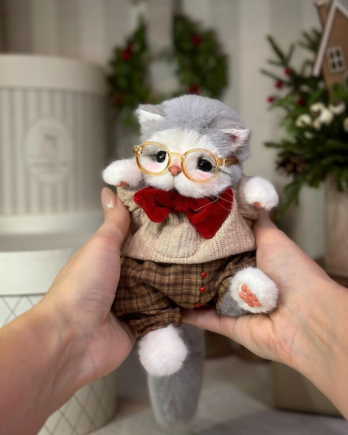 Handmade whimsical plush toy cat with red bow tie and glasses, held between two hands.
