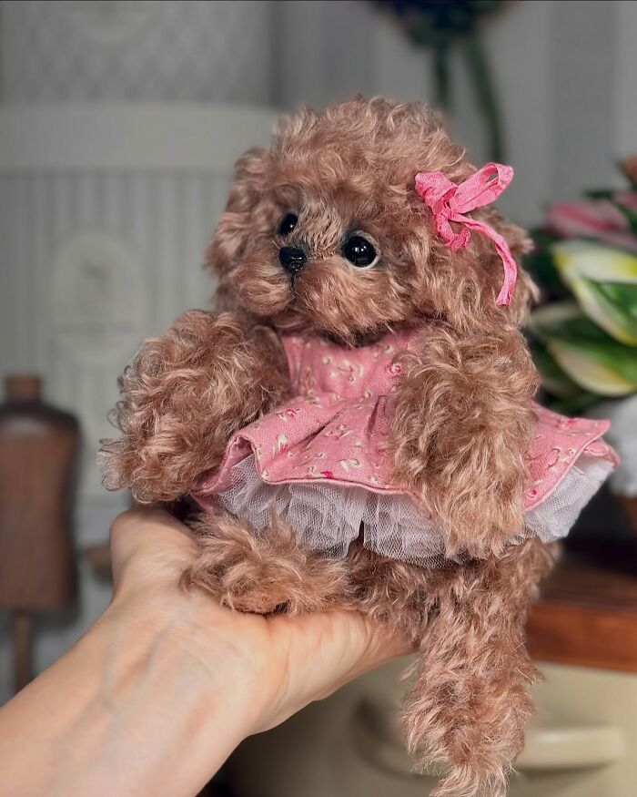 Handmade whimsical plush toy dog wearing a pink dress and bow, held in a person's hand indoors.