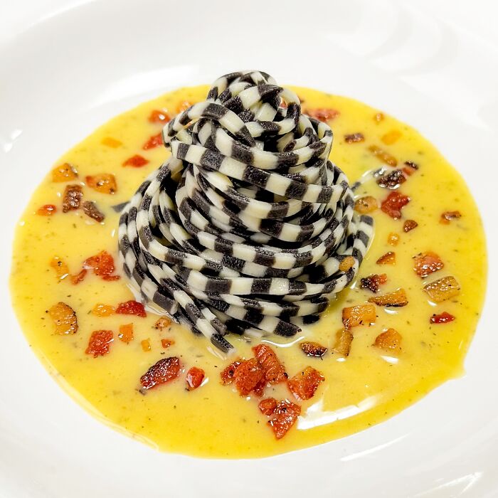 Black and white handcrafted pasta with colorful patterns, served on a creamy sauce with diced garnish.