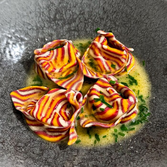 Colorful handcrafted pasta with red and yellow stripes, garnished in a light sauce in a dark bowl.