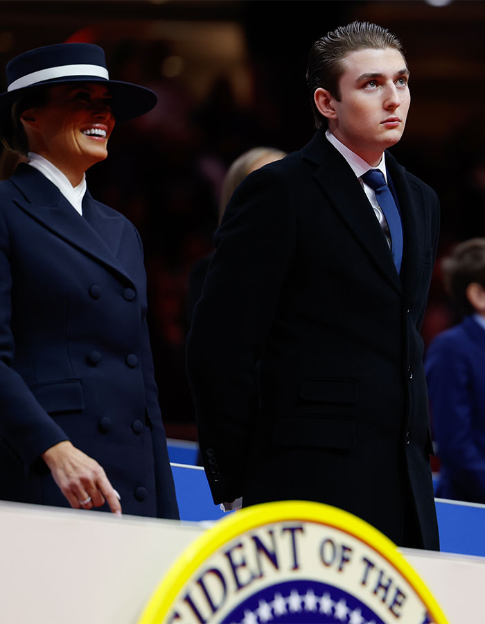 A young man in a dark suit stands beside a smiling woman in a hat, both observing an event with a presidential seal nearby. A young man in a dark suit stands beside a smiling woman in a hat, both observing an event with a presidential seal nearby.
