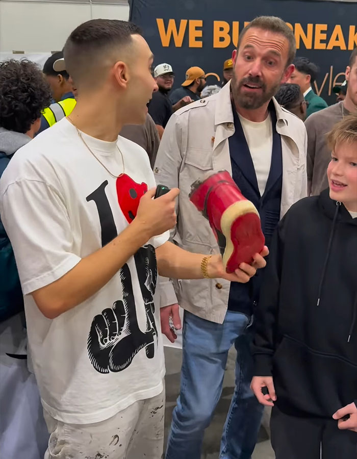 Man holding a sneaker interacts with Affleck at a bustling event, highlighting rich kids working regular shop jobs. Man holding a sneaker interacts with Affleck at a bustling event, highlighting rich kids working regular shop jobs.