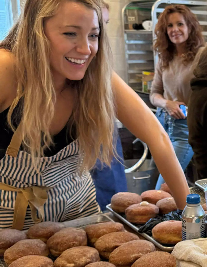 A woman in a striped apron smiling at a donut shop counter with trays of donuts. A woman in a striped apron smiling at a donut shop counter with trays of donuts.