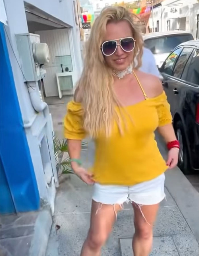 Blonde woman in sunglasses and yellow top, walks confidently on a sunny street, blue and white buildings around her. Blonde woman in sunglasses and yellow top, walks confidently on a sunny street, blue and white buildings around her.