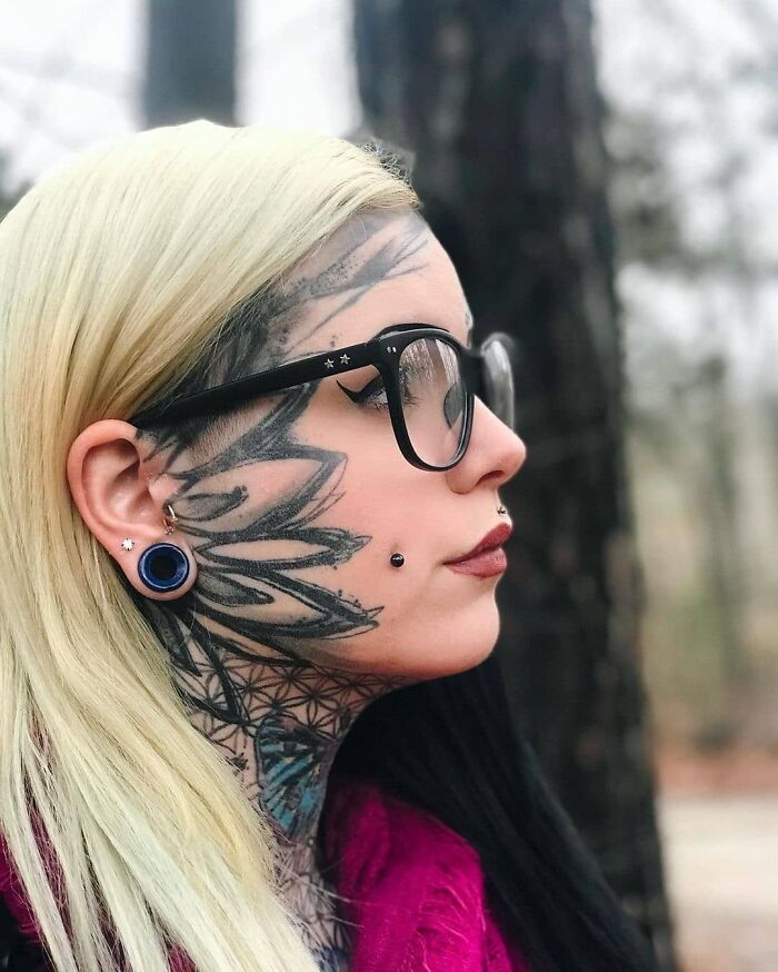 Person with elaborate face tattoos, glasses, and piercings in an outdoor setting, showcasing unique body art.