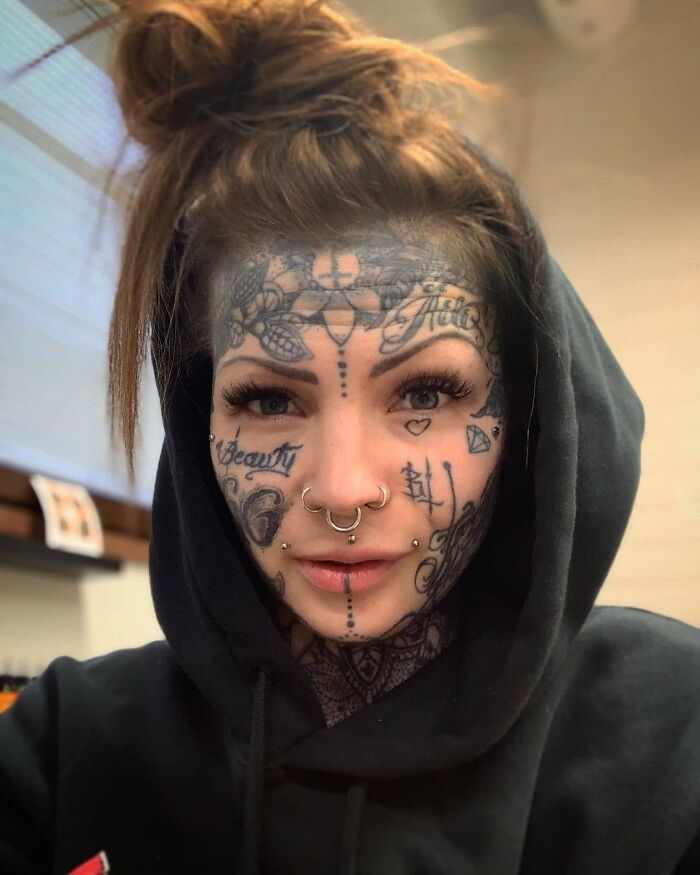 Person with face tattoos and piercings wearing a black hoodie, showcasing unique body art tattoos.