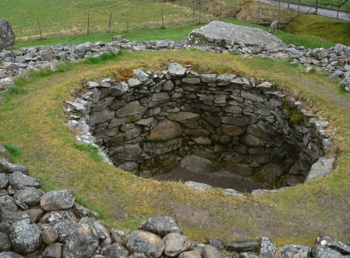 Ancient man-made stone structure with circular design surrounded by grass and scattered rocks in an outdoor setting