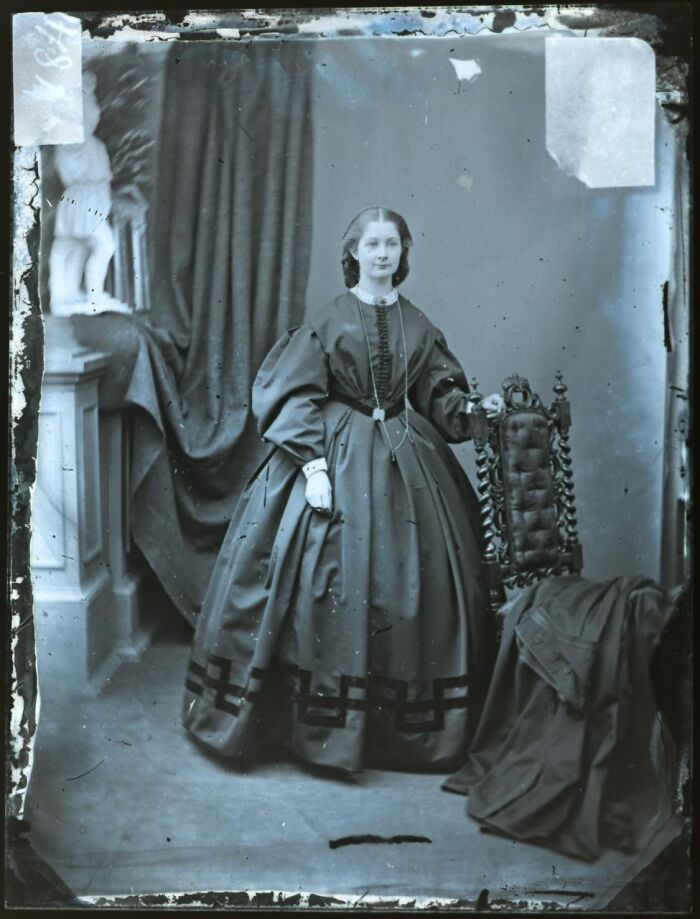 Victorian era woman in elaborate dress standing next to ornate chair in rare photograph rescued from glass negatives.