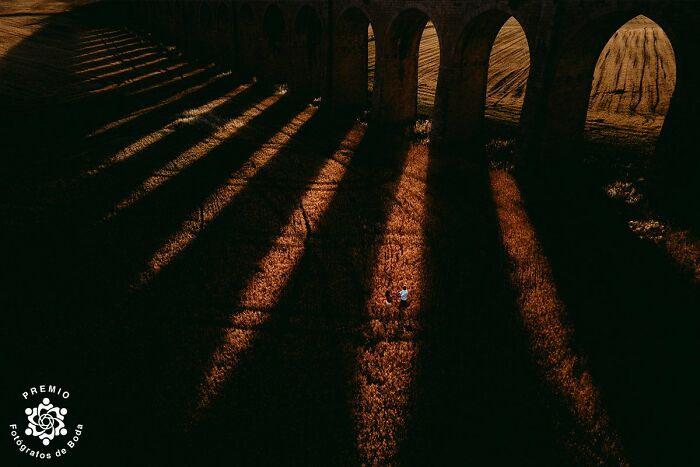 A couple stands under dramatic shadows cast by an old stone bridge, captured at the 2025 Premios FDB Wedding Awards.