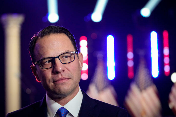 Man in glasses in front of bright lights, serious expression, blurred flags in background; related to Pennsylvania Governor's Mansion. Man in glasses in front of bright lights, serious expression, blurred flags in background; related to Pennsylvania Governor's Mansion.