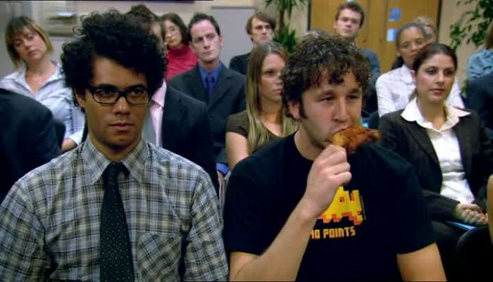 Two men sitting in a meeting, one in a checkered shirt and glasses, the other eating, surrounded by an audience.