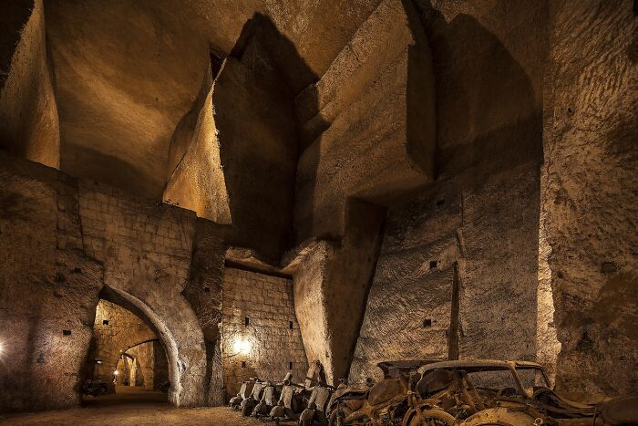 Eerie underground city with large stone arches and old vehicles, dimly lit and mysterious ambiance.