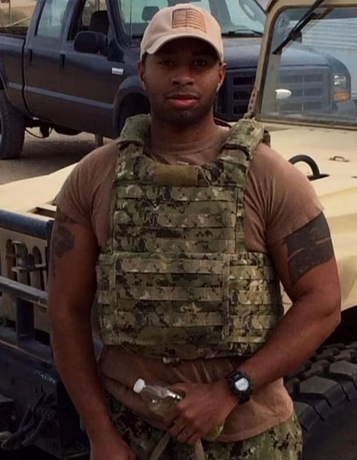 Former Navy SEAL in tactical gear holding a bottle, standing in front of a vehicle. Former Navy SEAL in tactical gear holding a bottle, standing in front of a vehicle.