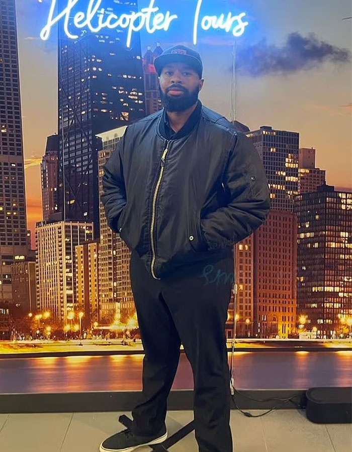 Man in front of cityscape backdrop under "Helicopter Tours" sign, wearing a black jacket and cap. Man in front of cityscape backdrop under "Helicopter Tours" sign, wearing a black jacket and cap.