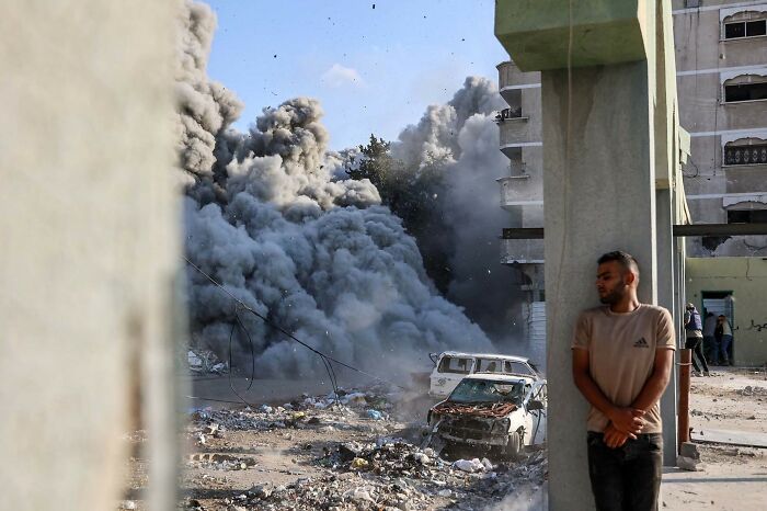 Man leans against a building as smoke billows from an explosion in a war-torn area, capturing powerful 2025 Istanbul Photo Awards moment.