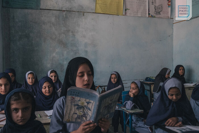 Girls in a classroom wearing blue uniforms, one reading aloud, from Istanbul Photo Awards.