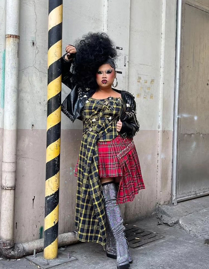 Drag Race alum in bold outfit with plaid patterns and metallic boots, standing by a striped pole outdoors. Drag Race alum in bold outfit with plaid patterns and metallic boots, standing by a striped pole outdoors.
