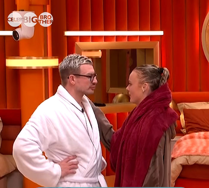 Two contestants share a moment in the Big Brother house, standing in a bright, orange-themed room. Two contestants share a moment in the Big Brother house, standing in a bright, orange-themed room.