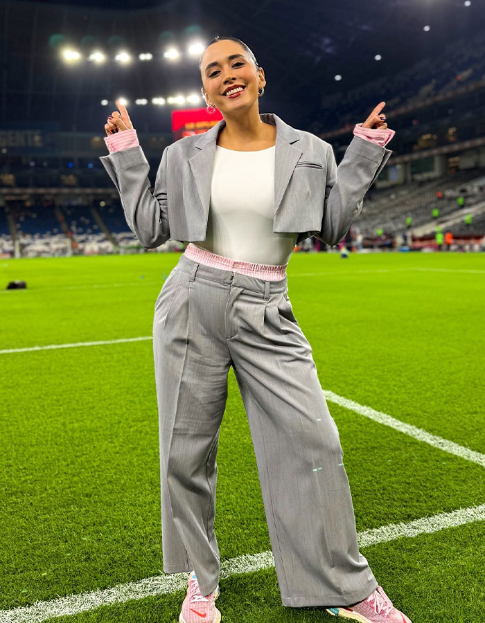 A female reporter in a grey suit smiles on a soccer field, highlighting media controversy over public reactions. A female reporter in a grey suit smiles on a soccer field, highlighting media controversy over public reactions.