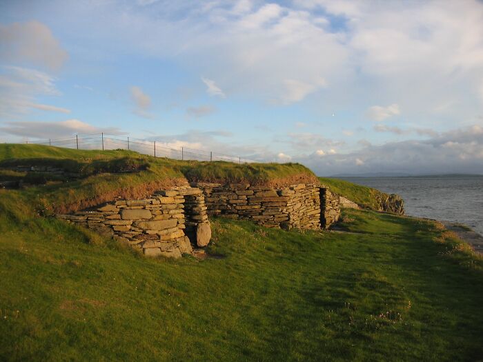 Ancient man-made stone structures built into grassy hillside near a coastline under a partly cloudy sky.
