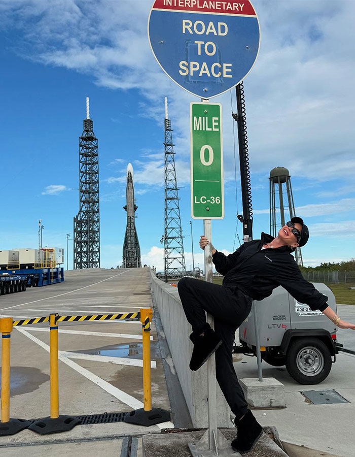 Person in black outfit posing under "Road to Space" sign at a space launch site, highlighting space mission theme. Person in black outfit posing under "Road to Space" sign at a space launch site, highlighting space mission theme.