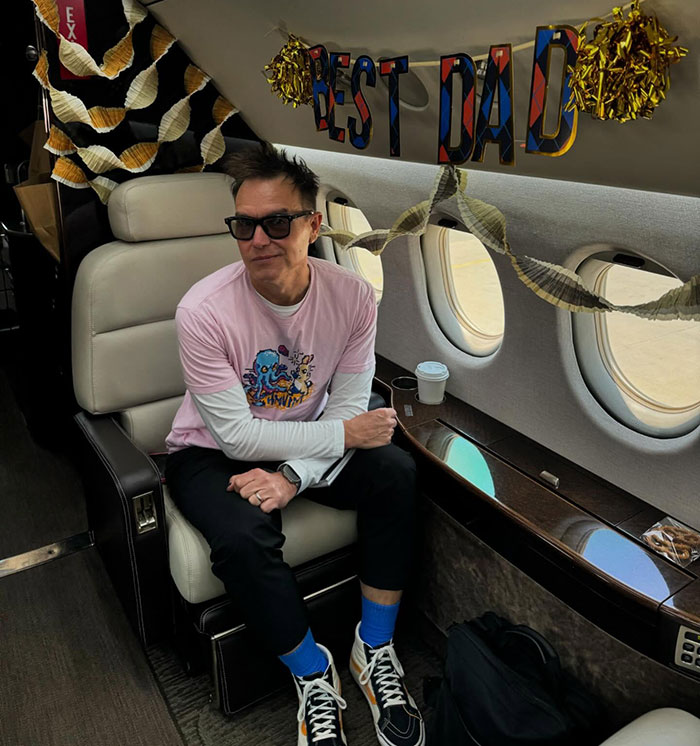 Mark Hoppus from Blink-182 sits in a private jet with "Best Dad" banner, wearing a pink shirt and sunglasses. Mark Hoppus from Blink-182 sits in a private jet with "Best Dad" banner, wearing a pink shirt and sunglasses.